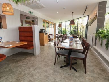 A dining area with wooden tables and chairs set for meals with large windows and plants at Lodge 3 in St Asaph