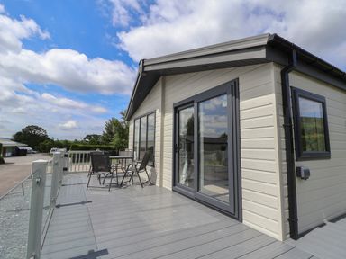An outdoor deck with a table and four chairs next to a beige building with large windows and sliding glass doors at Lodge 3 in St Asaph