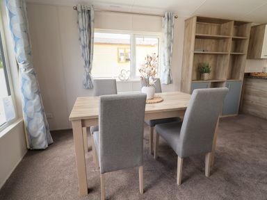 A dining area with a wooden table and four grey chairs near windows with blue patterned curtains and a wooden shelving unit at Lodge 3 in St Asaph