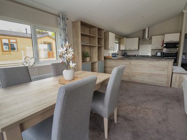 A dining area with a wooden table and grey chairs next to a kitchen with wooden cabinets and a countertop at Lodge 3 in St Asaph