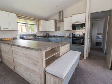 A kitchen with an L-shaped countertop, built-in oven and microwave, a bench with a cushion, and a doorway leading to a bedroom at Lodge 3 in St Asaph