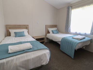 A bedroom with two single beds with blue bed runners and pillows next to a window at Lodge 3 in St Asaph