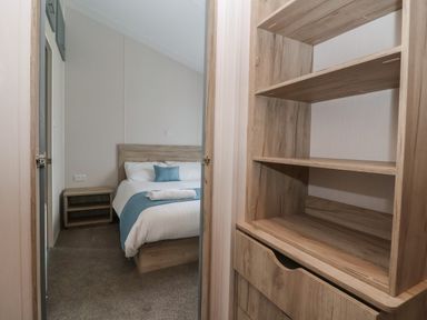 A bedroom with a wooden bed and nightstand seen through a doorway with wooden shelves on the right at Lodge 3 in St Asaph