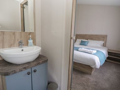 A bedroom with a made bed and blue decorative items seen through a partially open door next to a bathroom sink with hand soap at Lodge 3 in St Asaph