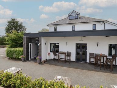 An outdoor seating area with wooden tables and chairs outside a white building with black windows and doors at Lodge 3 in St Asaph