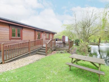 A wooden cabin with a ramp and stairs next to a pond with trees and a picnic table on grass at Kingfisher Lodge in Badwell Ash