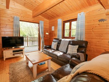A wooden living room with two dark leather sofas a coffee table television and windows with blue check curtains at Kingfisher Lodge in Badwell Ash