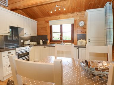 A kitchen with white cabinets a stove a window with a blind and a dining table with chairs and a decorative bowl at Kingfisher Lodge in Badwell Ash