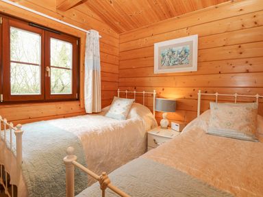 A wooden bedroom with two single beds a window with curtains and a nightstand with a lamp and decoration at Kingfisher Lodge in Badwell Ash