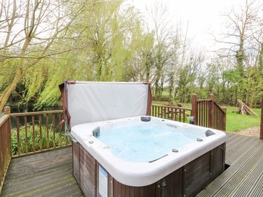 An outdoor hot tub with water and a cover on a wooden deck surrounded by trees and grass at Kingfisher Lodge in Badwell Ash