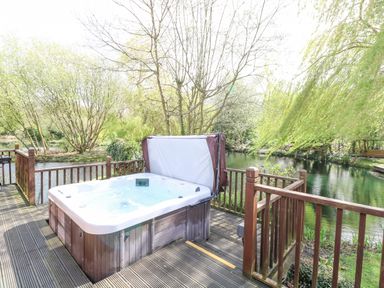 An outdoor hot tub on a wooden deck overlooking a river surrounded by trees at Kingfisher Lodge in Badwell Ash