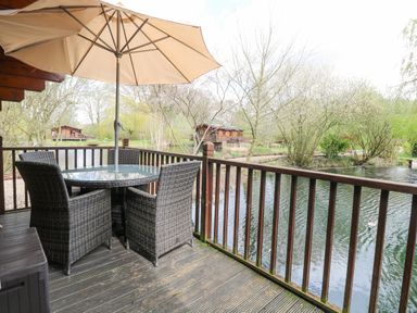 A deck with a round glass table with four wicker chairs and an umbrella overlooking a pond and trees at Kingfisher Lodge in Badwell Ash