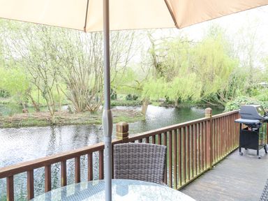 A deck with a glass table and umbrella beside a river with trees and a grill on the side at Kingfisher Lodge in Badwell Ash