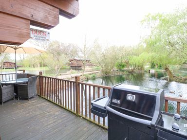 A deck with a barbecue grill and patio furniture overlooking a pond with trees and wooden cabins at Kingfisher Lodge in Badwell Ash