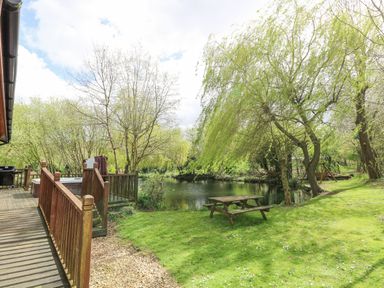 A wooden deck with railing, a hot tub, and a barbeque next to a grassy area with a picnic table by a pond surrounded by trees at Kingfisher Lodge in Badwell Ash