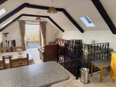 An attic living and dining area with exposed beams a dining table chairs a brown leather sofa and armchair a television telescope and balcony doors at Crogal Stable in New Quay