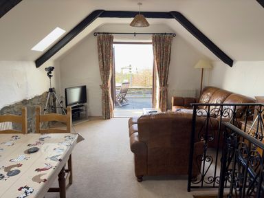 A living room with leather sofas a television on a stand a dining table with a tablecloth and an open door leading to a patio at Crogal Stable in New Quay