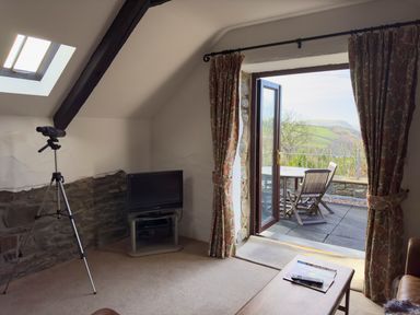 A living room with a television and telescope next to an open door leading to an outdoor patio with a table and chairs at Crogal Stable in New Quay
