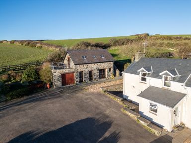 Two buildings with stone and white walls and a large paved area with grassy hills in the background at Crogal Stable in New Quay