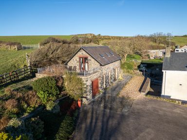 A stone building with a slate roof and balcony surrounded by grass and bushes at Crogal Stable in New Quay