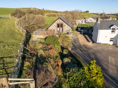 A stone house with a garden and fenced area next to a driveway and a white house in a rural setting at Crogal Stable in New Quay