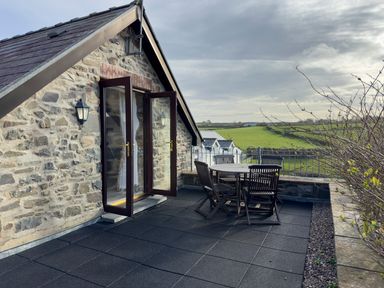 A stone building with open glass doors leading to a roof terrace with a wooden table and chairs overlooking green fields at Crogal Stable in New Quay