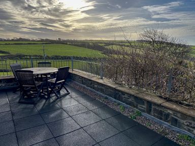 An outdoor patio with a round wooden table and four wooden chairs overlooking green fields and a cloudy sky at Crogal Stable in New Quay