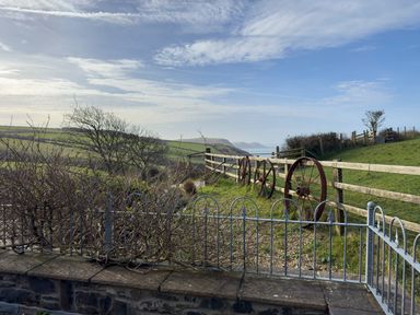 A garden area with a metal fence and wooden fence with wheels alongside a grassy hill overlooking distant hills and sea at Crogal Stable in New Quay