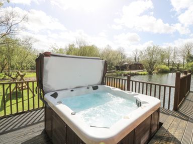 An outdoor hot tub on a wooden deck overlooking a pond with trees and a small cabin in the background at Wagtail Lodge in Badwell Ash