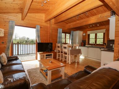 A wooden interior with a seating area with leather sofas and a coffee table a dining area with a table and chairs and a kitchen with white cabinets and windows at Wagtail Lodge in Badwell Ash