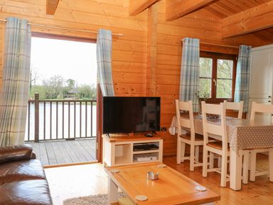 A wooden living room with a leather sofa a television on a stand a dining table with chairs and an open door to a balcony at Wagtail Lodge in Badwell Ash