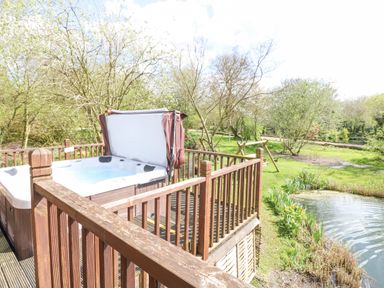 An outdoor wooden deck with a hot tub next to a pond and trees at Wagtail Lodge in Badwell Ash