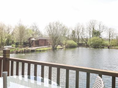 A wooden railing overlooking a pond with trees and a cabin in the background at Wagtail Lodge in Badwell Ash