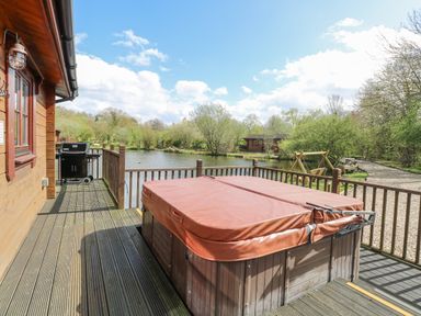 A deck with a covered hot tub and a barbecue overlooking a pond with trees and a wooden cabin at Woodpecker Lodge in Badwell Ash