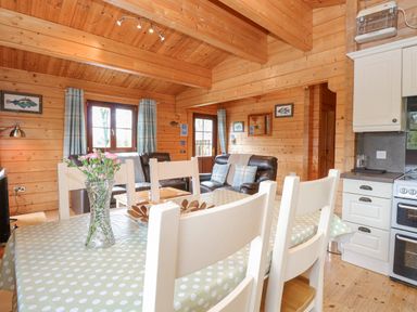 A wooden interior room with a dining table with chairs a vase with flowers leather sofas windows and kitchen appliances at Woodpecker Lodge in Badwell Ash