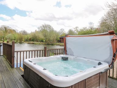 An outdoor hot tub on a wooden deck next to a lake with trees in the background at Woodpecker Lodge in Badwell Ash