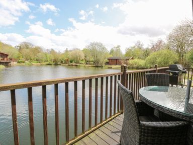A wooden deck with a glass table and wicker chairs overlooking a lake with cabins and trees at Woodpecker Lodge in Badwell Ash
