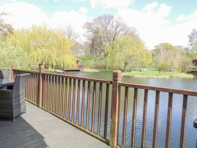 A wooden balcony with a table and chairs overlooking a pond with trees at Woodpecker Lodge in Badwell Ash