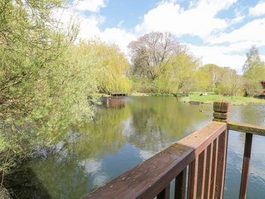 A wooden railing overlooking a pond surrounded by trees and grass at Woodpecker Lodge in Badwell Ash