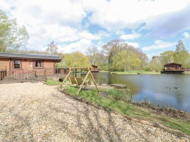 A wooden cabin by a lakeside with a wooden swing gravel path and trees at Woodpecker Lodge in Badwell Ash