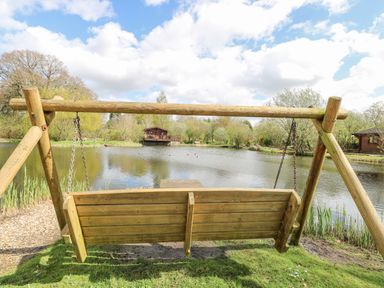 A wooden swing overlooking a pond with trees and wooden cabins at Woodpecker Lodge in Badwell Ash