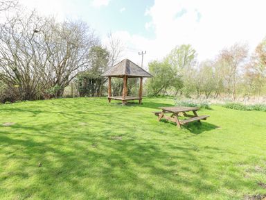 A grassy outdoor area with a wooden picnic table and a gazebo surrounded by trees at Woodpecker Lodge in Badwell Ash