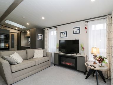 A living room with a beige sofa a tv on a stand with an electric fireplace and a side table with a lamp and flowers at Tiggywinkles in Chipping Norton
