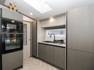 A kitchen with black microwave and oven stacked by a wooden door and a sink with black faucet under a window at Tiggywinkles in Chipping Norton