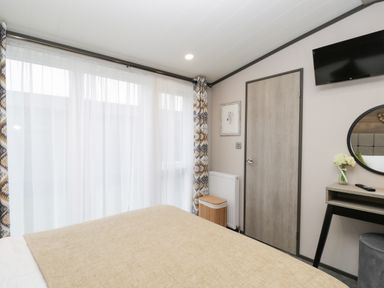 A bedroom with a bed beige blanket a door a wall-mounted television a dressing table with a mirror and flowers in a vase at Tiggywinkles in Chipping Norton