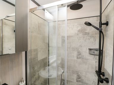 A shower area with glass sliding doors a black shower head and hand shower a wall-mounted wire basket and tiled walls at Tiggywinkles in Chipping Norton