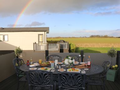 An outdoor dining table with food and drinks on a patio with a rainbow and house in the background at Tiggywinkles in Chipping Norton