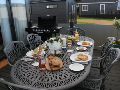 An outdoor dining table with plates of food bread bottles and glasses next to a barbecue grill at Tiggywinkles in Chipping Norton