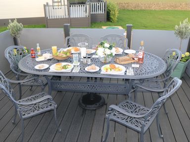 A metal outdoor dining table with six chairs set with plates of food drinks and flowers on a deck at Tiggywinkles Chipping Norton