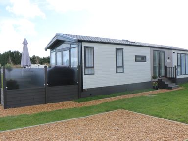 An exterior view of a modern single-story mobile home with a fenced patio and gravel pathway at Tiggywinkles in Chipping Norton